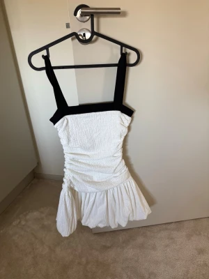 Vit kort klänning med svarta axelband - Trendig vit kort klänning med breda svarta axelband och smockad överdel. Klänningen har rynkade detaljer i midjan och en volangig kjol som ger en cool och ungdomlig look. Perfekt för varma dagar och sommarens alla tillfällen.