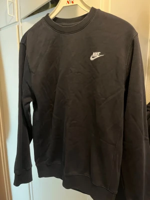 Svart crewneck sweatshirt från Nike - Svart sweatshirt från Nike med klassisk crewneck och vit broderad logga på bröstet. Tröjan har långa ärmar, ribbade muddar och är gjord i mjukt bomullsmaterial. Enkel och stilren design som passar till det mesta.