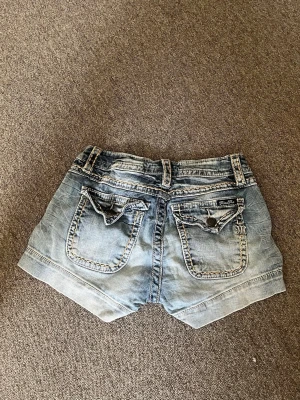 Blå jeansshorts från Miss Me - Snygga blå jeansshorts från Miss Me med midwaist passform. Shortsen har slitna detaljer, två bakfickor med lock och knapp samt dekorativa sömmar. Perfekta för varma dagar och en avslappnad stil.