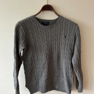 Grå kabelstickad tröja Polo Ralph Lauren - Snygg grå kabelstickad tröja från Polo Ralph Lauren med rund halsring. Tröjan är i storleken Small och är perfekt för alla årstider speciellt sommar kvällar.