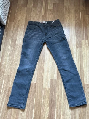 Grå jeans med raka ben - Säljer ett par grå jeans med klassisk femficksdesign och raka ben. Jeansen har subtila slitningar vid låren och är tillverkade i ett mjukt denim-material. Perfekt för dig som gillar en enkel och stilren look.