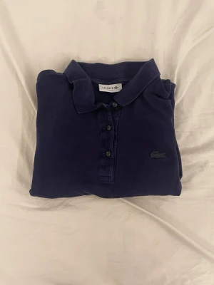 Mörkblå pikétröja från Lacoste - Klassisk mörkblå pikétröja från Lacoste med krage, knappar framtill och den ikoniska krokodilloggan broderad på bröstet. Tillverkad i bomull för en bekväm och stilren look. Perfekt till jeans eller shorts.
