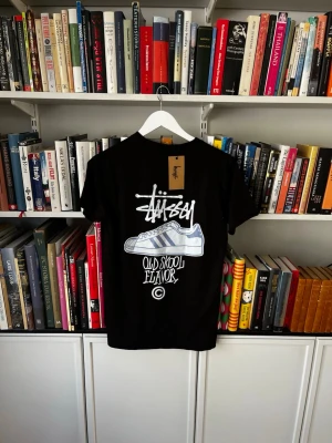 Svart Stüssy t-shirt med tryck - Svart t-shirt från Stüssy med stort grafiskt tryck på ryggen som visar en sneaker och texten 'Old Skool Flavor'. Framtill finns Stüssy-loggan i vitt. Klassisk passform och korta ärmar. Perfekt för dig som gillar streetwear.