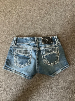 Blå jeansshorts från Miss Me - Snygga blå jeansshorts från Miss Me med dekorativa nitar och broderade detaljer på bakfickorna. Shortsen har låg midja, dubbla knappar och tydliga kontrastsömmar. Perfekta för sommaren och ger en cool, avslappnad vibe.
