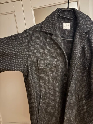 Grå overshirt från John Henric - Stilren grå overshirt/vårjacka från John Henric med diskret mönster. Klassisk krage, två bröstfickor med knappar och två sidofickor. Perfekt lager-på-lager-plagg i ett mjukt och lite tjockare material som ger en snygg och avslappnad look.