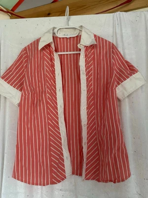  Randig skjorta med kort ärm - Säljer en rosa skjorta med vita vertikala ränder från Indian Rose. Skjortan har korta ärmar, vit krage och vita detaljer längs knappraden och ärmsluten. Perfekt för dig som gillar en avslappnad men ändå snygg stil.