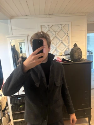 Mörkgrå overshirt från Zara - Säljer en stilren lite tjockare mörkgrå overshirt med hög krage och knäppning framtill. Overshirten har två bröstfickor med lock och är tillverkad i ett tjockt, ull-liknande material som ger en snygg och modern look. Perfekt för lager-på-lager och passar till många outfits. Storlek L men passar bättre på M