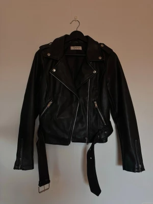 Svart skinnjacka från Nelly  - Cool svart skinnjacka köpt på Nelly med klassisk bikerstil, sned dragkedja, silverfärgade detaljer och bälte i midjan. Jackan har två fickor med dragkedja och axelklaffar som ger en edgy look. Perfekt för dig som vill ha en tidlös och trendig jacka.