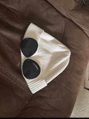 Vit ribbad mössa från C.P. Company - Säljer en vit ribbad mössa från C.P. Company med de ikoniska svarta glasögon-linserna på framsidan. Mössan är tillverkad i mjukt stickat material och har uppvikt kant. Perfekt för dig som vill ha en cool och trendig look.