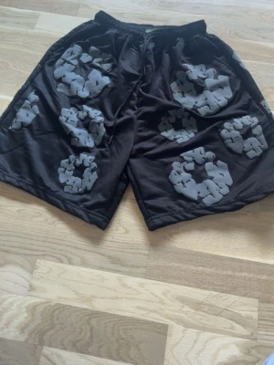 Svarta shorts från Denim Tears - Säljer ett par svarta shorts från Denim Tears med unikt grått blommönster över hela plagget. Shortsen har elastisk midja och är tillverkade i ett mjukt material som känns skönt mot huden. Perfekta för en avslappnad och trendig stil. Passar både som har storlek M-L