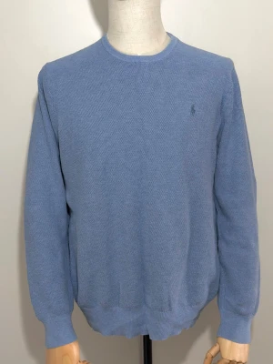 Blå stickad tröja från Ralph Lauren - Snygg blå stickad tröja från Ralph Lauren med rund halsringning och diskret broderad logga på bröstet. Tröjan har lång ärm och ribbade muddar vid ärmslut och nederkant. Perfekt för dig som gillar klassisk och stilren design.