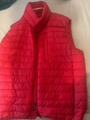 Röd dunväst från Esprit - Snygg röd dunväst från Esprit med hög krage och quiltad design. Västen har dragkedja framtill och en bröstficka med dragkedja. Perfekt för lager på lager och ger en sportig look till din outfit.