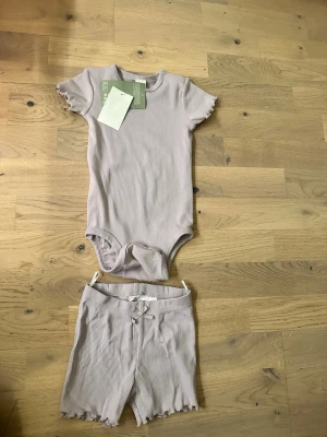 Ljuslila ribbat bodyset med shorts - Säljer ett sött set med ribbad body och matchande shorts i ljuslila. Bodyn har korta ärmar och volangkant, medan shortsen har liten rosett och samma volangdetalj. Båda delar är mjuka och stretchiga, perfekta för en avslappnad look.
