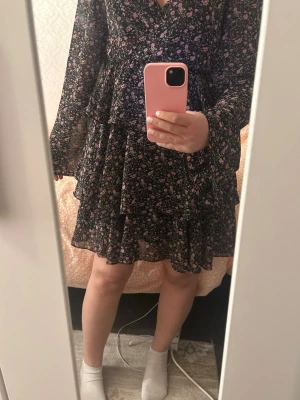 Svart blommig kortklänning från NA-KD - Supersöt svart volangig kortklänning från NA-KD med rosa och vita små blommor. Klänningen har volangdetaljer, v-ringning och långa ärmar. Perfekt för sommarkvällar!! Använd en kväll💓💓