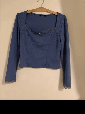 Blå ribbad topp med vit rosett - Supersöt blå långärmad topp med ribbad struktur, spetskant vid halsringning och ärmslut samt en liten vit rosett framtill. Croppad passform och fyrkantig urringning.