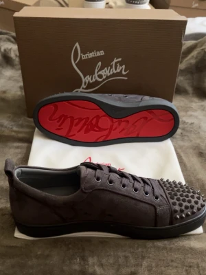 Christian Louboutin grå spikes sneakers - Gråa sneakers från Christian Louboutin i mocka med ikoniska nitar framtill och klassisk röd sula. Lågt skaft och svarta snören ger en edgy vibe. Perfekta för dig som vill sticka ut med exklusiv streetstyle.
