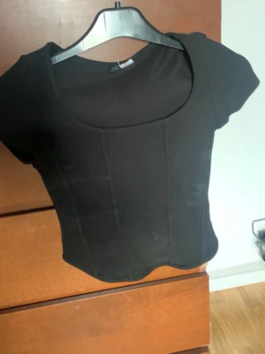 Svart croppad korsettopp - Snygg svart croppad topp med korsett-inspirerad design och korta ärmar. Toppen har markerade sömmar framtill och en rundad urringning. Perfekt att styla med höga jeans eller kjol för en trendig look.