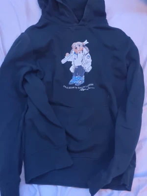 Svart hoodie Polo Ralph Lauren XL - Svart hoodie från Polo Ralph Lauren med ikoniska Polo Bear-trycket på bröstet. Klassisk huva och ribbade muddar. Perfekt för dig som gillar streetstyle och vill ha något bekvämt men ändå snyggt. Materialet känns mjukt och skönt mot huden.
