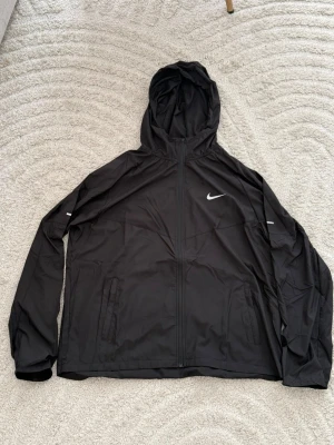 Svart Nike vindjacka med huva - Svart vindjacka från Nike med huva och dragkedja framtill. Jackan har två sidofickor med dragkedja och diskreta reflexdetaljer på ärmarna. Materialet är lätt och smidigt, perfekt för att skydda mot vind. Nike-loggan är tryckt på bröstet.