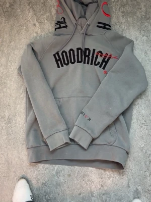 Grå hoodie från Hoodrich med tryck - Säljer en grå hoodie från Hoodrich med svart och rött tryck på bröstet och detaljer på huvan. Hoodien har en stor ficka framtill, ribbade muddar och justerbar huva med snören. Perfekt för dig som gillar streetwear och vill ha något bekvämt och snyggt.