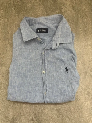 Blå linneskjorta från Polo Ralph Lauren - Säljer en snygg ljusblå skjorta från Polo Ralph Lauren i linne. Skjortan har klassisk krage, knappar framtill och det ikoniska Polo-logotypen broderad på bröstet. Perfekt för dig som gillar stilrena plagg med premiumkänsla.