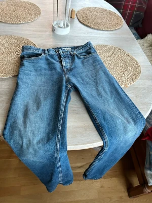 Tiger - Blå raka jeans med slitningar - Säljer ett par klassiska blå jeans med raka ben och snygga slitningar på framsidan. Jeansen har fem fickor, knappgylf och är tillverkade i bomull. Perfekt för dig som gillar en avslappnad och tidlös look.