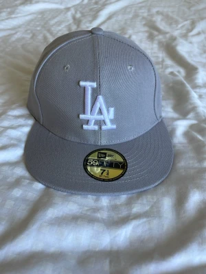 Grå LA New Era 59FIFTY keps - Snygg grå keps från New Era med LA-logga broderad i vitt framtill. Modellen är 59FIFTY med rak skärm och klassisk passform. Kepsen har små ventilationshål och New Era-logga på sidan. Perfekt för dig som gillar streetwear och sportig stil.