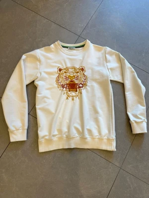 Vit sweatshirt med tiger från Kenzo - Säljer en vit sweatshirt från Kenzo med det ikoniska broderade tigermotivet och texten 'Kenzo Paris' på bröstet. Tröjan har rund halsringning, långa ärmar och ribbade muddar. Perfekt för dig som gillar streetwear och vill sticka ut.