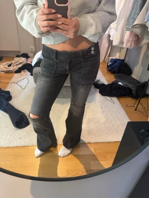 Gina Young Bootcut jeans med hål - Säljer ett par grå bootcut jeans med låg midja och hål på knät och fickan. Jeansen har dubbla knappar i midjan och stora fickor fram. Perfekta för en avslappnad och trendig look. Använder aldrig!
