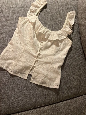 Vit broderad topp med volang - Söt vit topp med broderade blommor och volangdetaljer vid axelbanden. Toppen har knäppning framtill och är ärmlös, perfekt för sommardagar.💕