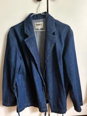 Blå jeansjacka från ONLY - Snygg blå jeansjacka/blazer från ONLY med klassisk krage och långa ärmar. Jackan har en avslappnad passform och knytband i midjan för en cool look. 