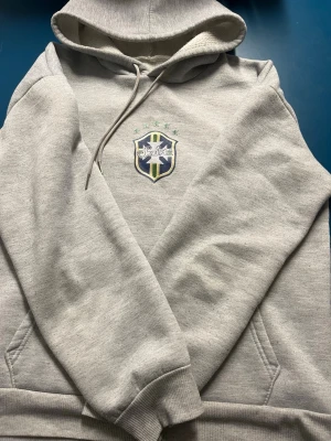 Brazil tracksuit  - Snygg grå tracksuit med Brasilien-emblem på bröstet och klassiska kängurufickor. Tröjan har huva med dragsko och ribbade muddar vid ärmar och nederkant. Perfekt för dig som gillar sportig stil och vill visa support för Brasilien. Mjuk och skön bomullsblandning.