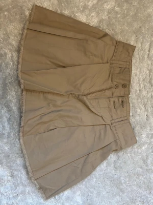 Beige kortkjol med rå kant - Snygg beige kortkjol med rå, fransad kant och två knappar framtill. Kjolen har bred linning och veckade detaljer som ger en cool och avslappnad look. Perfekt för dig som gillar en enkel men trendig stil.