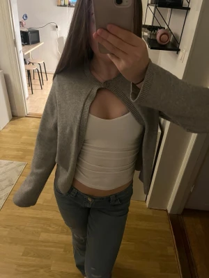 Grå cropped kofta med knapp - Säljer en grå cropped kofta med en knapp framtill. Koftan har långa ärmar och är i ett mjukt, stickat material. Perfekt att slänga över ett linne eller t-shirt för en chill look.