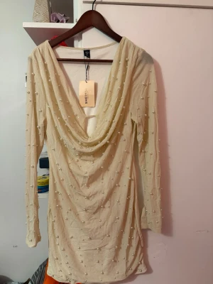 Beige klänning med pärlor från SHEIN - Trendig beige kortklänning från SHEIN med långa ärmar och dekorativa pärlor över hela tyget. Klänningen har en snygg draperad öppen rygg och är tillverkad i ett lätt syntetmaterial. Perfekt för dig som vill sticka ut med en unik detalj.