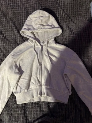 Ljusgrå croppad hoodie med dragkedja - Säljer en ljusgrå croppad hoodie med dragkedja framtill och huva med dragsnören. Hoodien har långa ärmar och bred mudd i midjan för en snygg passform. Perfekt för en avslappnad och trendig look.