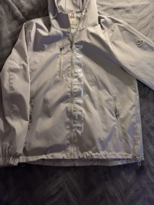 Beige vindjacka från Moncler - Snygg beige vindjacka från Moncler med stor logga längs dragkedjan framtill. Jackan har huva, två sidofickor med dragkedja och Moncler-märke på ärmen. Tillverkad i lätt och tåligt material, perfekt för blåsiga dagar.