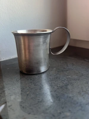 Äktasilvermugg med handtag - En stilren mugg i silver med rund form och ett brett, räfflat handtag. Muggen har en blank yta och passar perfekt för både varma och kalla drycker. Enkel och klassisk design som funkar i många sammanhang. Äkta silver 830  50gm