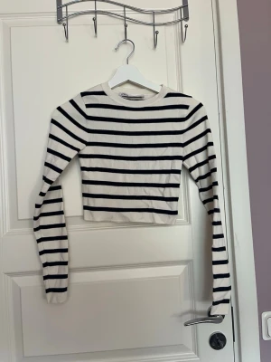 Randig croppad topp från Zara - Superfin croppad långärmad topp från Zara med svartvita ränder. Den har rund halsringning och är i ett mjukt, stretchigt material som sitter snyggt på kroppen. Perfekt att styla med jeans eller kjol för en trendig look.