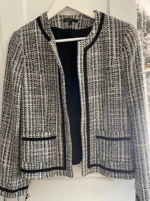 Tweedjacka från KappAhl svartvit - Snygg svartvit tweedjacka från KappAhl i storlek 38. Jackan har raka linjer, två framfickor och svarta kantband. Materialet är vävt i en klassisk tweedstruktur och jackan är helfodrad för extra komfort. Det går att stänga till med två haspar fram 