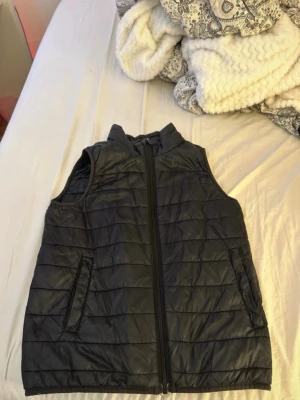 Svart quiltad dunväst med dragkedja - Snygg svart dunväst med quiltat mönster och hel dragkedja framtill. Västen har två sidofickor med dragkedja och står upp lite i kragen för extra stil. Perfekt att slänga över en hoodie eller tröja för en chill look.