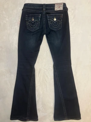 True Religion mörkblå joey low rise flare jeans - Säljer ett par mörkblå low rise joey flare jeans från True Religion med kontrastsömmar och ikoniska fickdetaljer bak. Jeansen har låg midja, klassisk femficksdesign och silverfärgade knappar. Perfekta för dig som gillar en avslappnad men ändå trendig look. Jeansen är i bra sick och användes ett par gånger