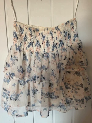 Blommig volangkjol i blått och vitt - Söt kortkjol med volanger och blommigt mönster i blått och vitt. Kjolen har en smockad midja för skön passform och är gjord i ett lätt, luftigt material som passar perfekt till varmare dagar. Perfekt för dig som gillar romantisk och feminin stil.