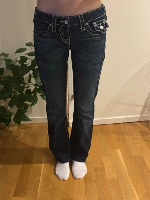 Mörkblå True Religion jeans - Säljer ett par mörkblå jeans från True Religion med vita sömmar. Jeansen har låg midja, raka ben och fickor fram och bak med diamanter på. Midjemått: 36cm.🩷Innerbenslängd: 85cm.🩷