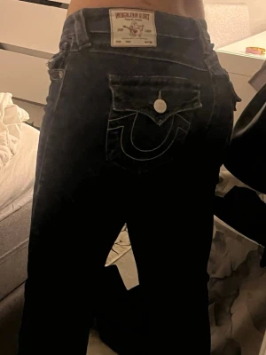 Mörkblå True Religion jeans - Säljer ett par mörkblå jeans från True Religion med klassisk kontrastsöm och ikonisk fickdetalj bak. Jeansen har låg midja, flare, och silverfärgade knappar. Materialet är stretchigt denim för skön passform. De är lite slitna längst nere vid fötterna men inget man märker ❤️