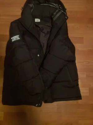 Svart jacka från Burberry - Säljer en svart dunväst från Burberry med huva och dragkedja. Västen har tryckknappar framtill, två sidofickor med dragkedja och logga på bröstet där det står 'Burberry London England'. Perfekt för lager på lager och riktigt snygg streetstil. Kan även tänka mig byteshandel