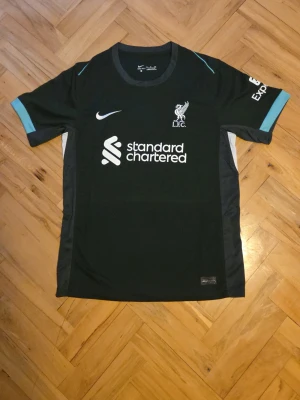 Liverpool Nike svart matchtröja - Snygg svart Liverpool FC matchtröja från Nike med vita och turkosa detaljer. Tröjan har klubbmärke och sponsortryck på bröstet samt korta ärmar med turkosa muddar. Perfekt för fotbollsträning eller att bära till vardags.