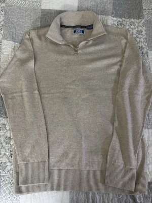 Beige stickad half zip från Jack & Jones - Beige stickad tröja från Jack & Jones med half zip och hög krage. Tröjan har ribbade muddar vid ärmslut och nederkant, perfekt för en clean och avslappnad stil. Passar till det mesta och är enkel att matcha.