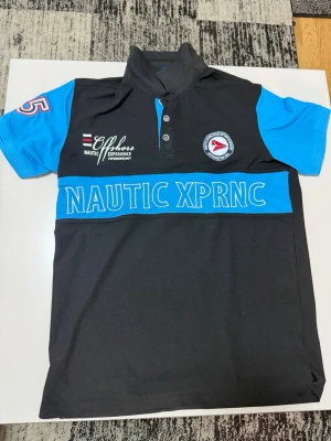 Svart och blå piké från Swedemount - Sportig pikétröja från Swedemount i svart och blått med tryck och broderade detaljer. Modellen har korta ärmar, knappslå och texten 'NAUTIC XPRNC' framtill. Perfekt för en avslappnad och aktiv stil.