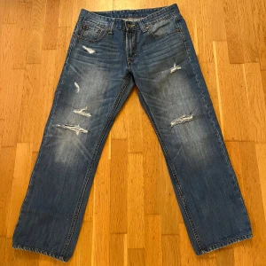 Vintage Levi's 504 jeans - Säljer ett par Vintage Levi's 504 jeans i blå tvätt med snygga slitningar och distressed detaljer på både fram- och baksida. Jeansen har rak passform, fem fickor och ikonisk röd Levi's-tag vid bakfickan. Perfekt för dig som gillar en avslappnad och cool stil.